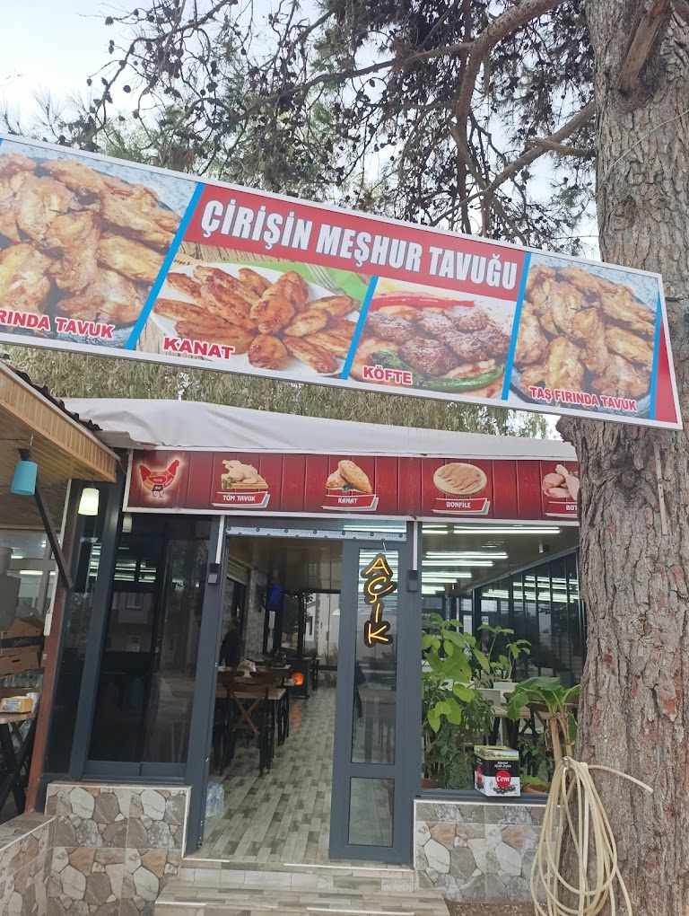 Çiriş Tavukçuluk restaurant entrance with a large outdoor sign displaying grilled chicken, wings, and köfte. The sign reads Cirişin Meşhur Tavuğu and includes menu items such as Kanat, Köfte, Taş Fırında Tavuk. The restaurant has a welcoming and casual atmosphere with tables inside, plants near the entrance, and a neon sign that says Açık indicating it is open. The setting is outdoors with trees and natural light, evoking a friendly and inviting mood.